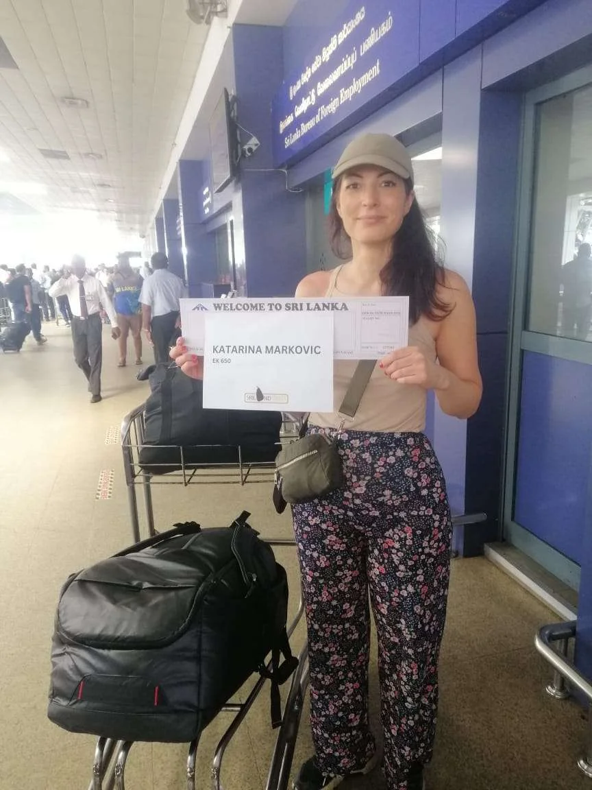 In an airport, a woman displays a sign saying "Trip to Sri Lanka," excitedly looking for someone.